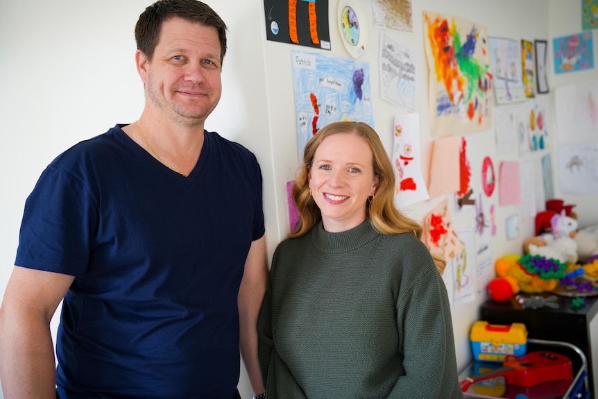 Warren and Kristen smile in front of a wall covered in their children's drawings.