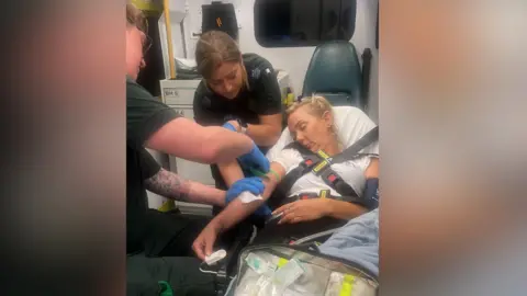 Michelle Sword's family newspaper is assisted by paramedics. She lies unconscious, strapped to an ambulance bed. Male and female paramedics assist her.