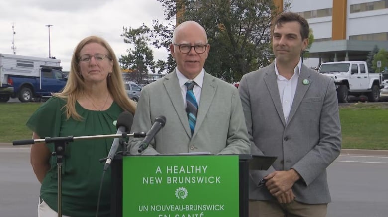 A man in a gray suit stands between a man and a woman outside at a microphone.