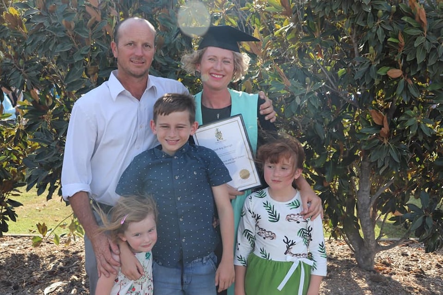 A woman in a graduation gown and hat stands with her family in front of a tree.