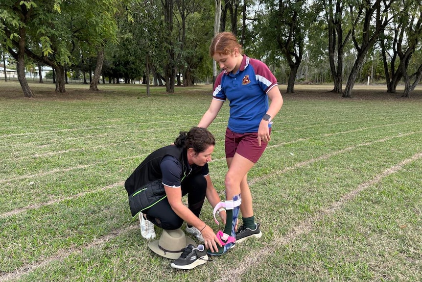 A woman helping a girl put on running shoes while standing on the grass of an oval.