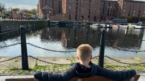 A boy with blond hair sits on a bench and looks at a dock with narrow boats on it.