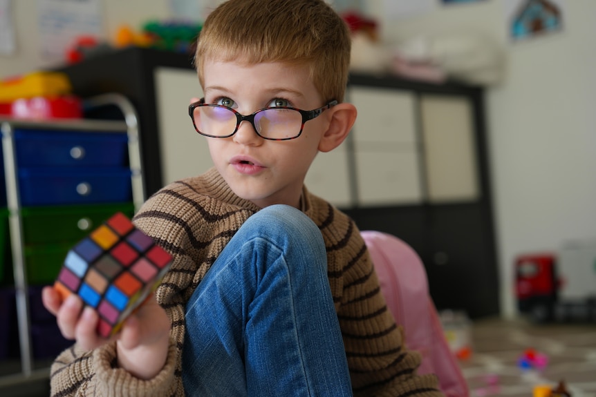 A cute kid with glasses looks up while playing with a Rubik's cube.